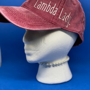 May include: A maroon baseball cap with a distressed, washed-out finish. The text "Lambda Lady." is embroidered in white, cursive-style lettering across the side panel of the cap. The cap features a curved brim and stitched eyelets on the crown.