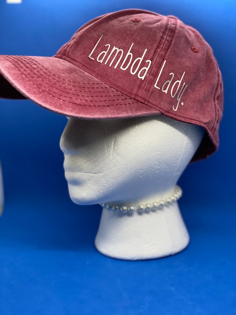 May include: A maroon baseball cap with a distressed, washed-out finish. The text "Lambda Lady." is embroidered in white, cursive-style lettering across the side panel of the cap. The cap features a curved brim and stitched eyelets on the crown.