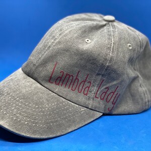 May include: A grey baseball cap with a distressed, washed-out texture. The cap features the text "Lambda Lady" printed in a dark red, sans-serif font across the front panel. It includes a curved brim with visible stitching and a button on top.