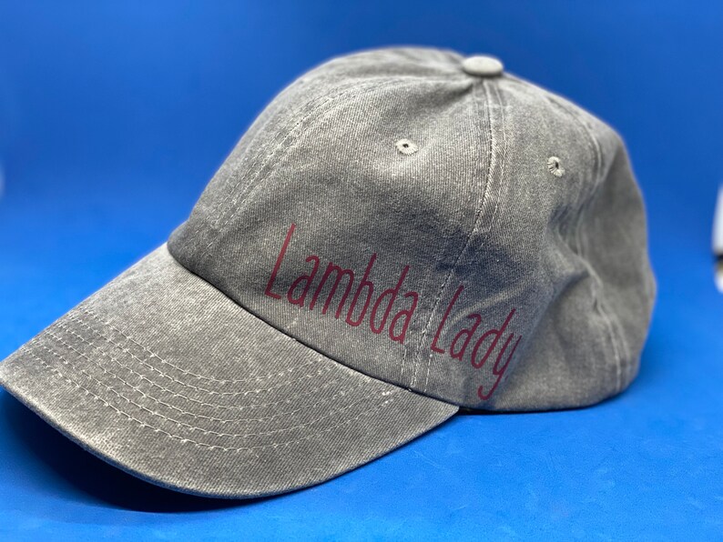 May include: A grey baseball cap with a distressed, washed-out texture. The cap features the text "Lambda Lady" printed in a dark red, sans-serif font across the front panel. It includes a curved brim with visible stitching and a button on top.