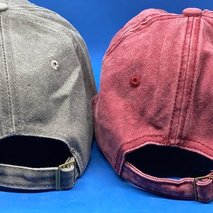 May include: Two baseball caps, one in a faded gray and one in a faded maroon, each featuring a six-panel construction with stitched eyelets and an adjustable fabric strap at the back secured with a metallic buckle for a customizable fit.