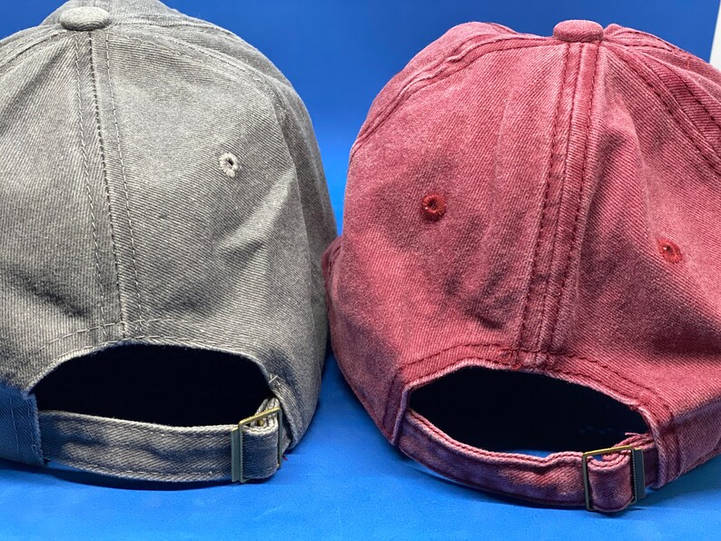 May include: Two baseball caps, one in a faded gray and one in a faded maroon, each featuring a six-panel construction with stitched eyelets and an adjustable fabric strap at the back secured with a metallic buckle for a customizable fit.