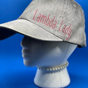 May include: A grey baseball cap with a distressed, washed-out texture. The text "Lambda Lady." is printed on the front in a dark pink, cursive-style font. The cap features a curved brim and stitched eyelets on the crown for ventilation.