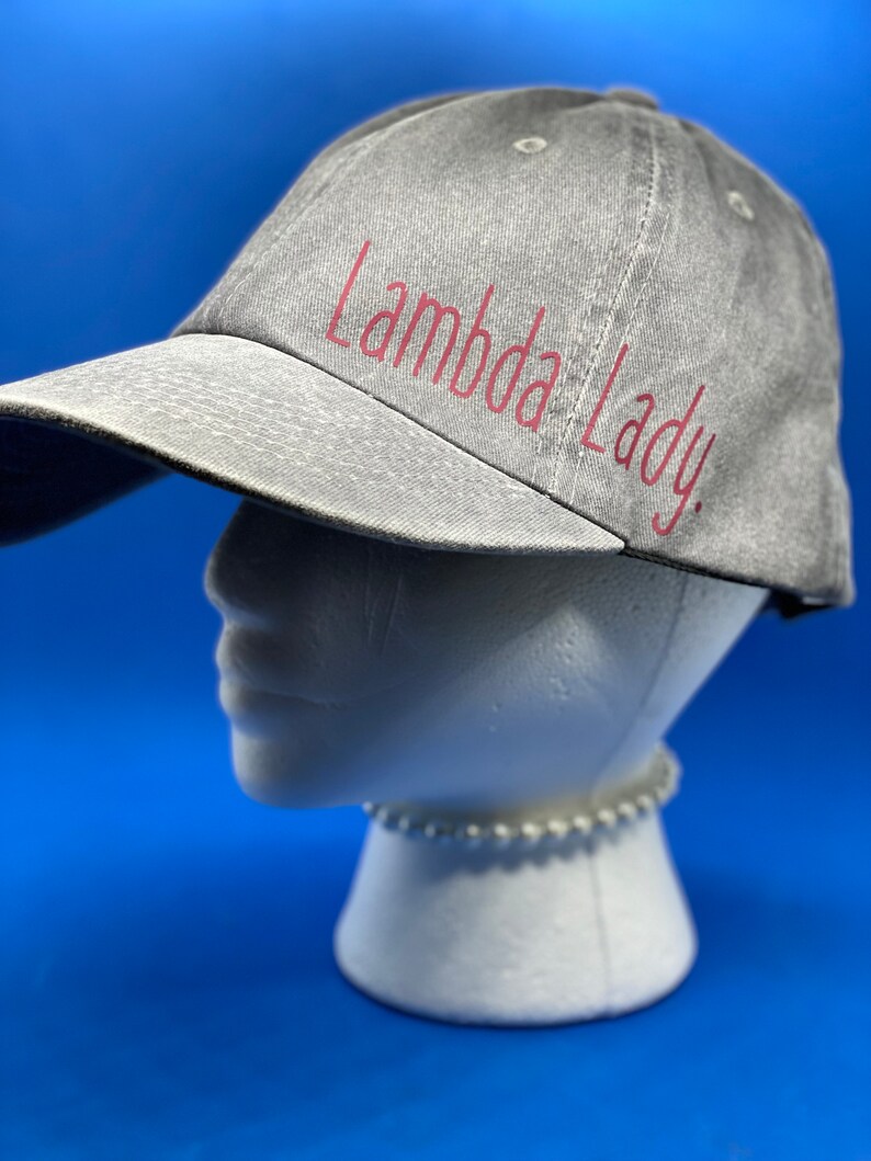 May include: A grey baseball cap with a distressed, washed-out texture. The text "Lambda Lady." is printed on the front in a dark pink, cursive-style font. The cap features a curved brim and stitched eyelets on the crown for ventilation.