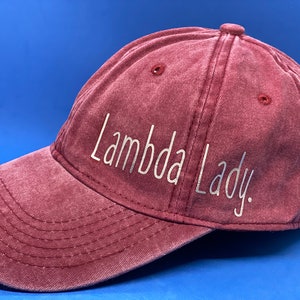 May include: A maroon baseball cap with a distressed, washed-out texture. The front panel features the text "Lambda Lady." printed in a clean, white, sans-serif font. The cap includes a curved brim with visible stitching and ventilation eyelets.