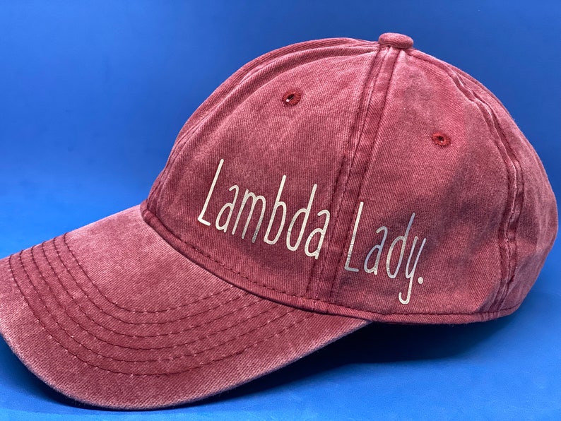May include: A maroon baseball cap with a distressed, washed-out texture. The front panel features the text "Lambda Lady." printed in a clean, white, sans-serif font. The cap includes a curved brim with visible stitching and ventilation eyelets.