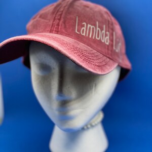 May include: A maroon baseball cap with a distressed, washed-out texture. The text "Lambda Lit" is embroidered in white thread on the front panel, positioned slightly to the right. The cap features a curved brim and visible stitching details.