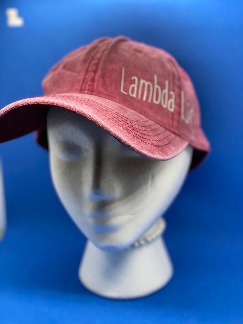 May include: A maroon baseball cap with a distressed, washed-out texture. The text "Lambda Lit" is embroidered in white thread on the front panel, positioned slightly to the right. The cap features a curved brim and visible stitching details.