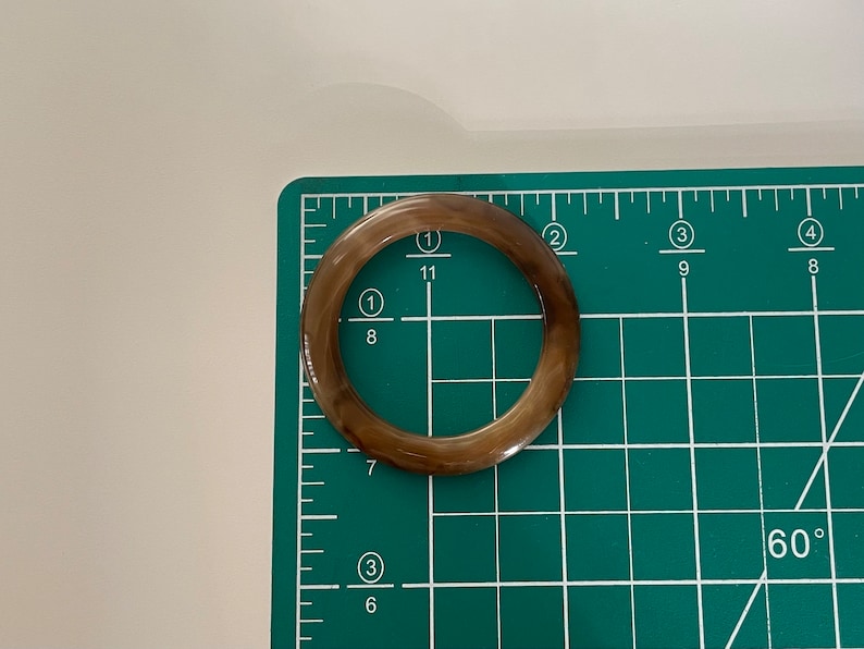 May include: A brown and white ring-shaped object with a smooth surface. The object is approximately 2 inches in diameter and is lying on a green cutting mat.