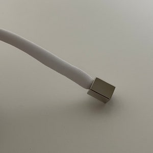 Puede incluir: Un cable blanco con un conector de metal plateado en el extremo.