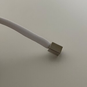 Puede incluir: Un cable blanco con un conector de metal plateado en el extremo.