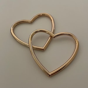 May include: Two gold heart-shaped hoops, one slightly larger than the other, are intertwined. The hoops are smooth and shiny, creating a simple yet elegant design.