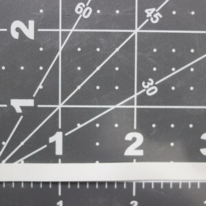 Może przedstawiać: Biały, prostokątny pasek materiału o wymiarach 2,5 cm x 7,6 cm, umieszczony na szarej macie do cięcia z białymi liniami siatki i numerami.