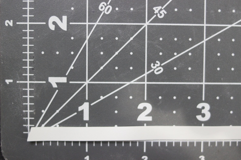 Może przedstawiać: Biały, prostokątny pasek materiału o wymiarach 2,5 cm x 7,6 cm, umieszczony na szarej macie do cięcia z białymi liniami siatki i numerami.