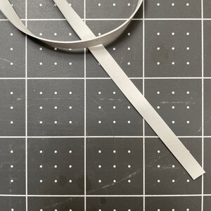 Może przedstawiać: Biała wstążka leży na szarej macie do cięcia z białymi kropkami. Wstążka jest złożona na p&oacute;ł, a końce skierowane są w przeciwne strony.