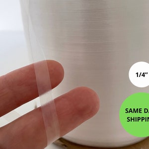以下が含まれることがあります： 幅1/4インチの透明なプラスチック製の梱包用テープのロール。テープは指で持ち、ロールから引き出されています。
