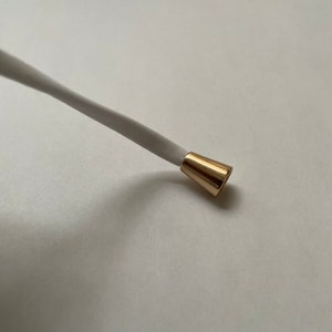 Puede incluir: Un tapón de cordón de metal dorado con un cordón blanco enhebrado.