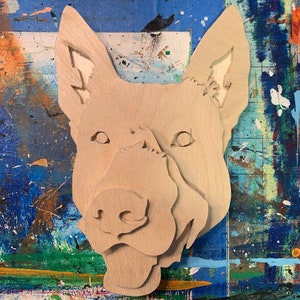 May include: A layered wooden art piece depicting a dog's head. The artwork is cut from light-colored wood, with details like the eyes, nose, and ears carved out. The dog's head is centered against a colorful, paint-splattered background.