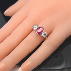 May include: A three-stone ring featuring a large oval-shaped ruby surrounded by two smaller round diamonds. The ring is set in a white metal band.