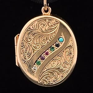 May include: Gold oval locket with an engraved floral design and a line of colorful gemstones. The locket is on a black ribbon.