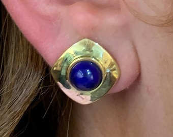 Könnte beinhalten: Goldene Ohrringe mit quadratischer Fassung und einem runden blauen Edelstein.