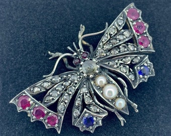 Könnte beinhalten: Eine silberne Schmetterlingsbrosche mit roten, weißen und blauen Edelsteinen. Der Schmetterling ist mit Diamanten und Perlen besetzt.