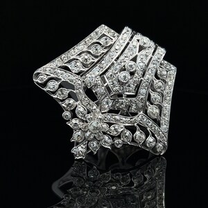 May include: A silver and diamond brooch with a geometric design. The brooch is shaped like a star and features a floral motif in the center.
