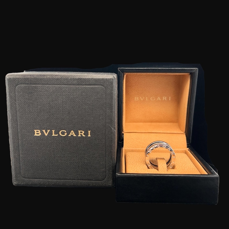 May include: A silver ring with a circular design, in a black and brown box with the word "BVLGARI" printed in gold on the box.