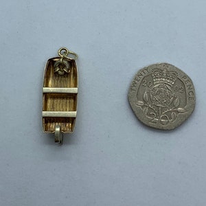 May include: A gold-toned charm in the shape of a small wooden rowboat. The charm has a loop at the top for attaching to a necklace or bracelet. The charm is about the size of a 20 pence coin.