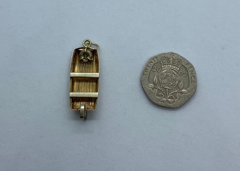 May include: A gold-toned charm in the shape of a small wooden rowboat. The charm has a loop at the top for attaching to a necklace or bracelet. The charm is about the size of a 20 pence coin.