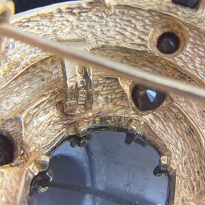 K&ouml;nnte beinhalten: Nahaufnahme der R&uuml;ckseite einer goldfarbenen Brosche mit einem schwarzen Stein und kleineren Steinen. Die Brosche hat einen Stift und einen Verschluss. Die R&uuml;ckseite der Brosche ist mit "925 C.C." gekennzeichnet.