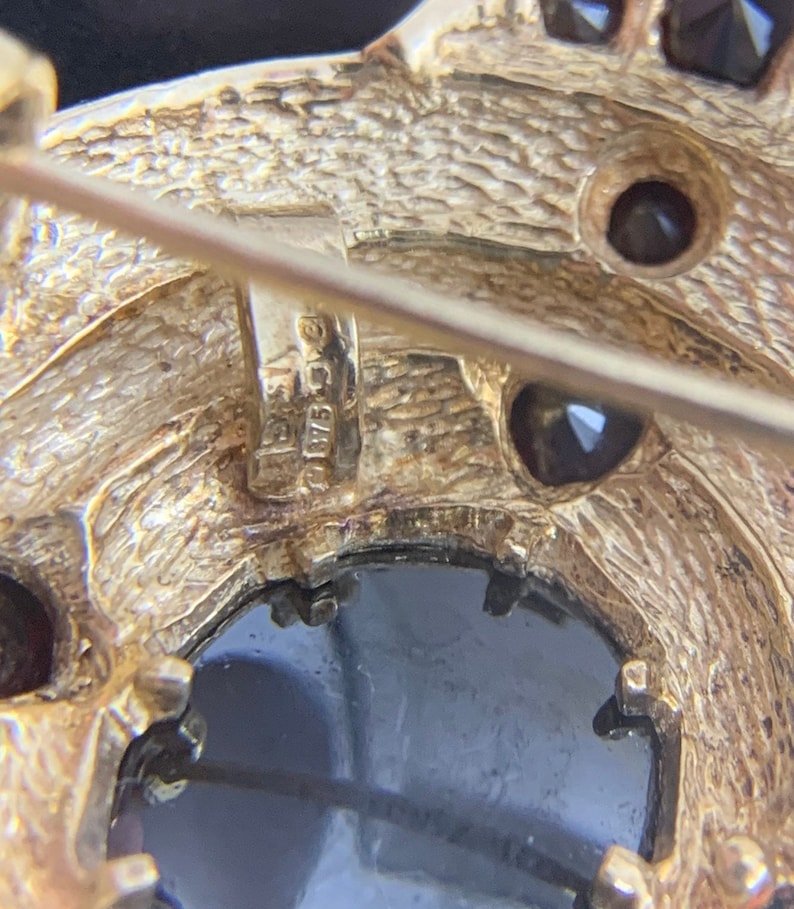 K&ouml;nnte beinhalten: Nahaufnahme der R&uuml;ckseite einer goldfarbenen Brosche mit einem schwarzen Stein und kleineren Steinen. Die Brosche hat einen Stift und einen Verschluss. Die R&uuml;ckseite der Brosche ist mit "925 C.C." gekennzeichnet.
