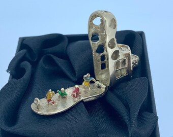 Puede incluir: Un anillo plateado articulado con diseño en forma de casa. El anillo se abre para revelar una pequeña plataforma con figuras en miniatura con atuendos coloridos. El anillo se presenta sobre una superficie de satén negro.