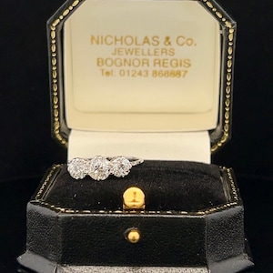 Puede incluir: Una caja de anillo blanca con un borde dorado y el texto "Nicholas & Co. Jewellers Bognor Regis Tel. 01243 868887" en su interior. Un anillo de diamantes de tres piedras est&aacute; sentado dentro de la caja.
