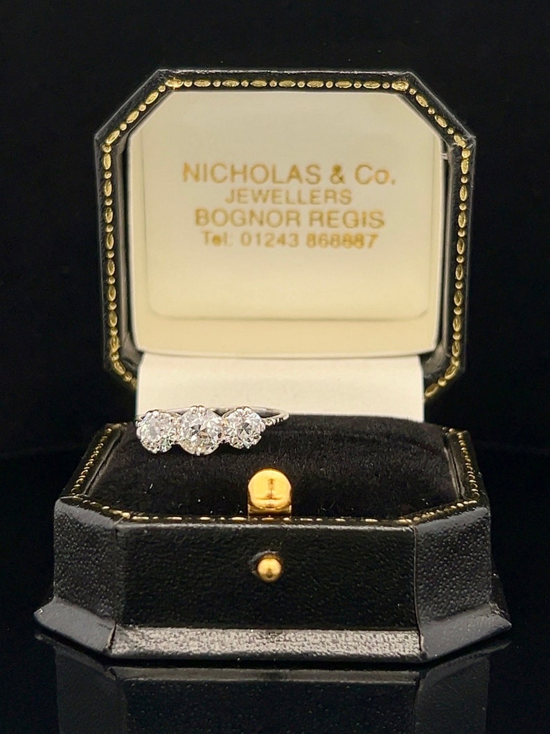 Puede incluir: Una caja de anillo blanca con un borde dorado y el texto "Nicholas & Co. Jewellers Bognor Regis Tel. 01243 868887" en su interior. Un anillo de diamantes de tres piedras est&aacute; sentado dentro de la caja.