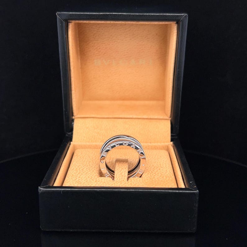 May include: A silver ring with multiple bands, each with a row of small diamonds, in a black box with a tan interior.
