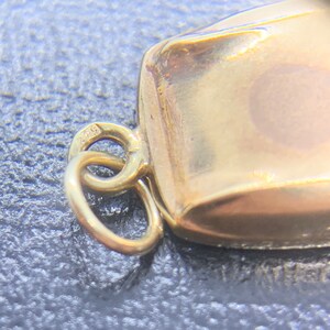 May include: A gold charm with a square shape and a loop for attaching to a necklace or bracelet. The charm has the number "975" stamped on it.