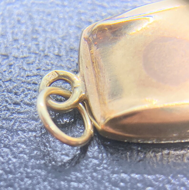 May include: A gold charm with a square shape and a loop for attaching to a necklace or bracelet. The charm has the number "975" stamped on it.