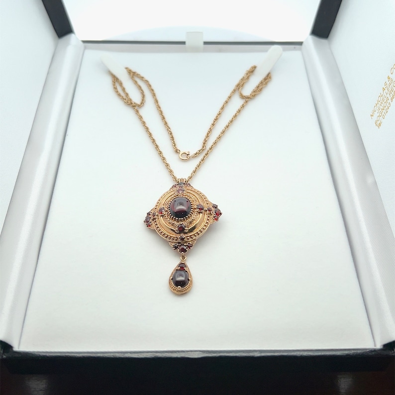 K&ouml;nnte beinhalten: Eine goldene Halskette mit einem Anh&auml;nger, der einen gro&szlig;en roten Edelstein zeigt, der von kleineren roten Edelsteinen umgeben ist. Der Anh&auml;nger hat die Form eines Quadrats mit abgerundeten Ecken und einen tropfenf&ouml;rmigen Anh&auml;nger, der daran h&auml;ngt.