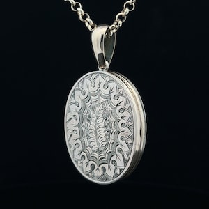 Op de afbeelding: Zilveren ovaal medaillon met een ingewikkeld gegraveerd ontwerp met een bloemenmotief. Het medaillon is bevestigd aan een zilveren ketting.