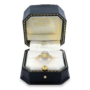 May include: A gold ring with a large oval-shaped center stone surrounded by smaller diamonds. The ring is in a black ring box with the text "NICHOLAS & Co. JEWELLERS BOGNOR REGIS Tel. 01243 864887" on the inside of the lid.