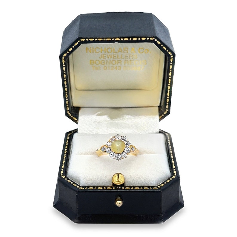 May include: A gold ring with a large oval-shaped center stone surrounded by smaller diamonds. The ring is in a black ring box with the text "NICHOLAS & Co. JEWELLERS BOGNOR REGIS Tel. 01243 864887" on the inside of the lid.