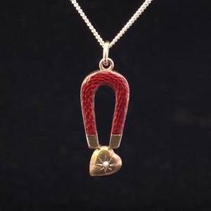 Puede incluir: Un collar con colgante de oro con un diseño de herradura de esmalte rojo y un dije en forma de corazón. El corazón tiene un diseño de estrella y una pequeña perla. La cadena del collar es plateada.
