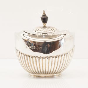 Peut inclure: Un récipient argenté avec couvercle, corps cannelé et un bouton décoratif. Le récipient est de forme arrondie avec un couvercle ajusté. Le bouton est brun foncé avec un accent doré. Le récipient est probablement en métal.