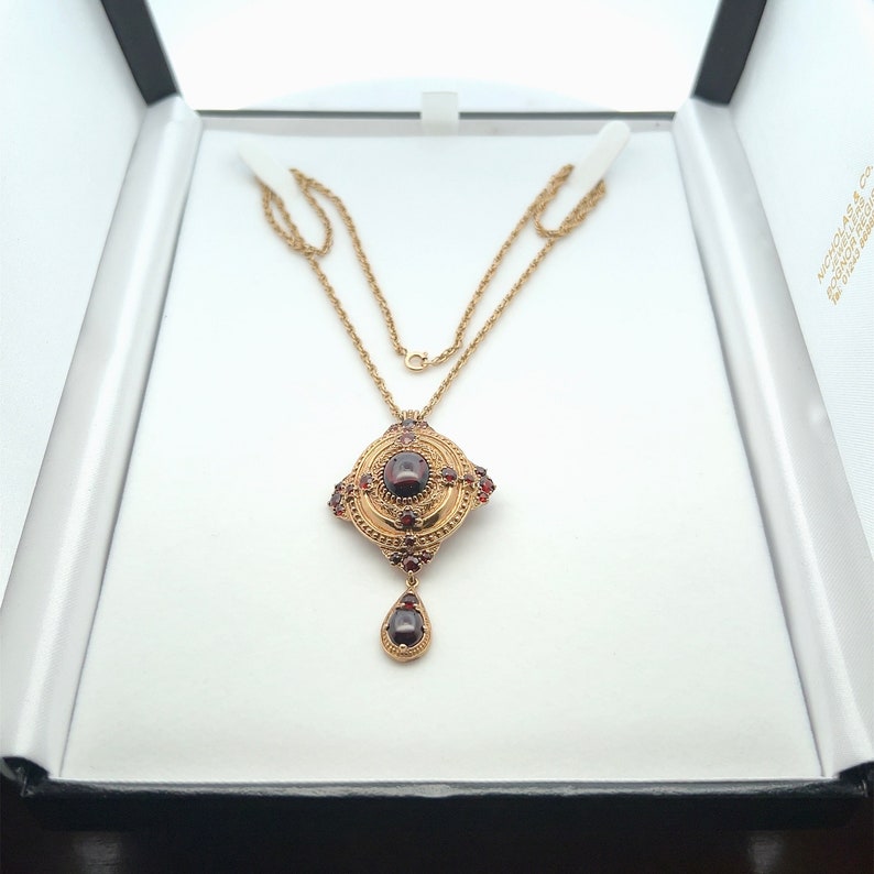 K&ouml;nnte beinhalten: Eine goldene Halskette mit einem Anh&auml;nger in quadratischer Form, der mit einem gro&szlig;en roten Edelstein verziert ist, der von kleineren roten Edelsteinen umgeben ist. Der Anh&auml;nger hat ein tropfenf&ouml;rmiges St&uuml;ck, das mit einem roten Edelstein an der Unterseite h&auml;ngt.