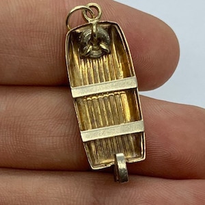 May include: Gold-toned metal charm shaped like a small boat with a rope detail. The charm is detailed with wood-like texture and a ring at the top for attaching to a necklace or bracelet.