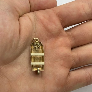 May include: A gold-colored charm in the shape of a small boat. The boat has a white stripe around the middle and a white stripe around the top. The charm is hanging from a loop at the top.