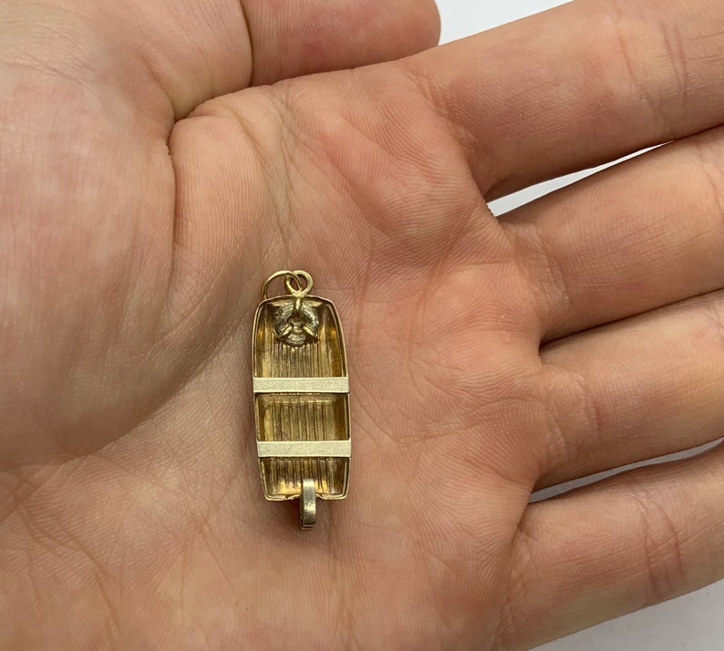 May include: A gold-colored charm in the shape of a small boat. The boat has a white stripe around the middle and a white stripe around the top. The charm is hanging from a loop at the top.
