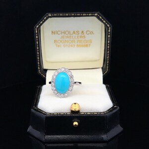 May include: A turquoise gemstone ring with a halo of diamonds set in white gold. The ring is in a black ring box with the name "Nicholas & Co. Jewellers Bognor Regis Tel 01243 860887" printed on the inside.