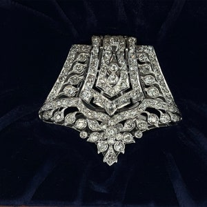 May include: A silver and diamond brooch with a geometric design. The brooch is shaped like a bell and has a large diamond in the center. The brooch is set on a dark blue velvet background.